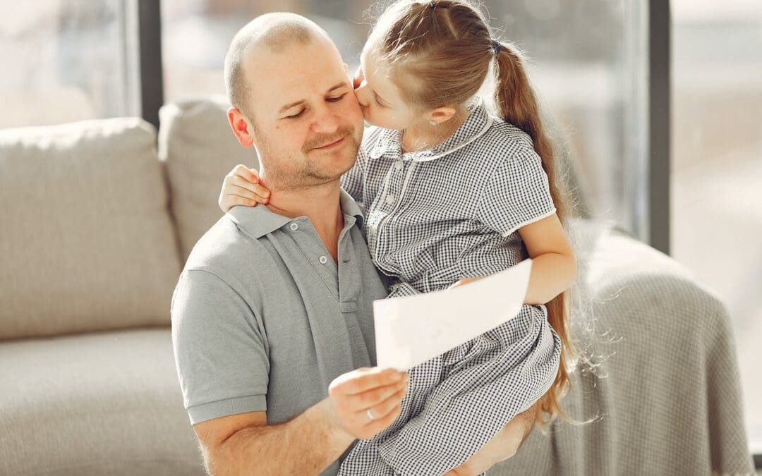 A dad with a low-cut beard holds his daughter, a girl with a long blonde ponytail, as she kisses his cheek. The man is absently reading a white paper while holding his daughter. They stand in front of a beige couch in a sunlit living room.